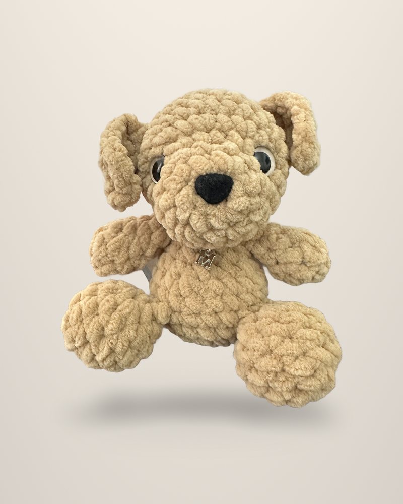 A crocheted puppy in toffee beige with long floppy ears and a small metal letter M charm, seated.