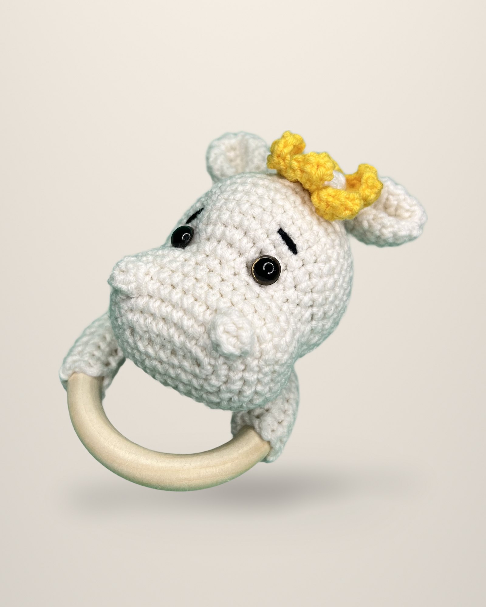 A crocheted ivory hippo head with a small yellow crown, attached to a pale ring.