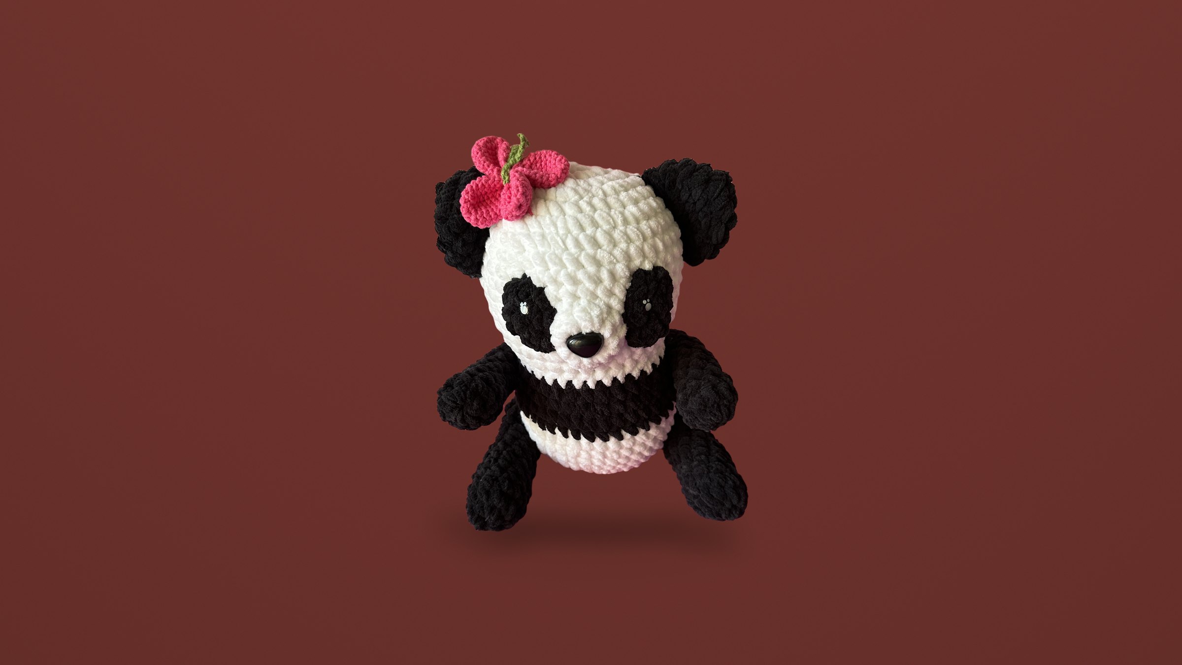 A crocheted panda in black and ivory with a pink flower on its head, on an ivory backdrop.