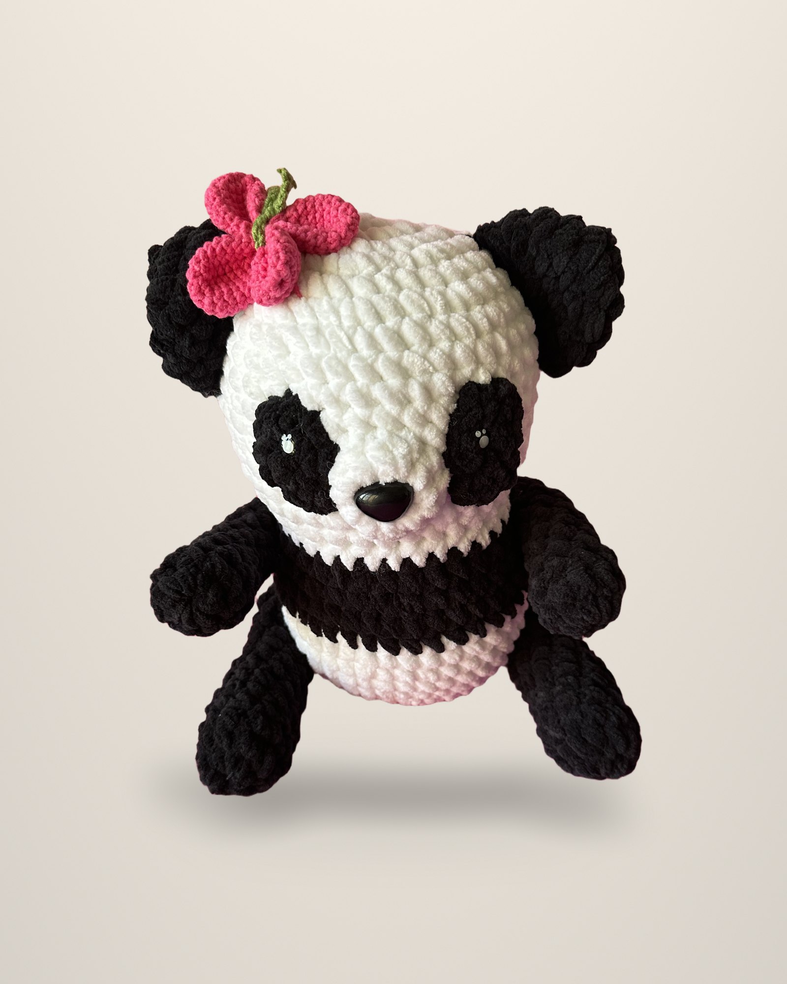A crocheted panda in black and ivory with a pink flower on its head, on an ivory backdrop.